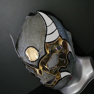 May include: A silver and gold lucha libre mask with white accents. The mask features a gold circular emblem on the side and a detailed design with sharp angles and curves. The mask is displayed on a mannequin head.