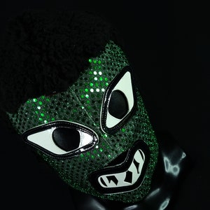 May include: A green wrestling mask with black and white details. The mask has a fierce expression with a large, toothy grin and two large eyes. The mask is covered in green sequins.