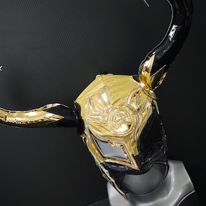 Peut inclure: Un masque de lutte doré et noir avec un design cornu. Le masque présente des ornements dorés, un emblème central et une base noire. Les cornes sont longues et incurvées, ajoutant à l'aspect dramatique. Le masque est présenté sur une tête de mannequin.