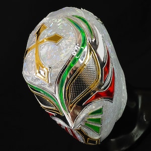 Op de afbeelding: Een wit worstelmasker met een zilver, groen en rood ontwerp. Het masker heeft een kruis op de voorkant en een zilveren en gouden rand.