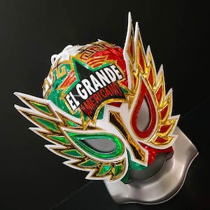 May include: A colorful wrestling mask with the text "EL GRANDE AMERICANO". The mask features a white base with green, red, and gold accents. The design includes stylized wings and geometric patterns, creating a bold and eye-catching appearance.