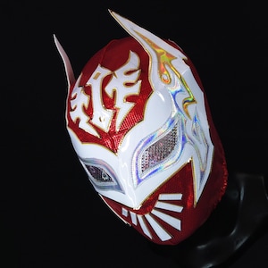 Op de afbeelding: Een rood en wit worstelmasker met een wit ontwerp en een gouden rand. Het masker heeft een wit ooggaas en een rood en wit patroon.