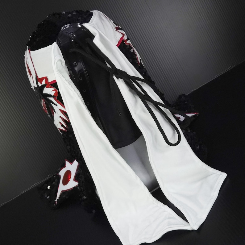 Puede incluir: Una m&aacute;scara de lucha libre profesional con un dise&ntilde;o blanco, negro y rojo. La m&aacute;scara presenta un ribete de lentejuelas negras, detalles rojos y un cord&oacute;n negro. La m&aacute;scara est&aacute; hecha de una combinaci&oacute;n de materiales.