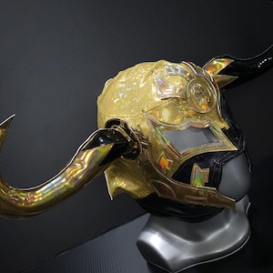 Peut inclure: Un masque de lucha libre doré et noir avec une surface dorée texturée et à paillettes. Le masque présente des cornes dorées, des accents irisés et un design détaillé autour des ouvertures des yeux. Le masque est exposé sur une tête de mannequin grise.