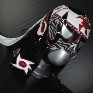 Puede incluir: Una m&aacute;scara de lucha libre con un dise&ntilde;o llamativo. La m&aacute;scara presenta un estampado de cebra en blanco y negro, acentuado con detalles rojos y blancos. Est&aacute; adornada con lentejuelas negras y una capa blanca.