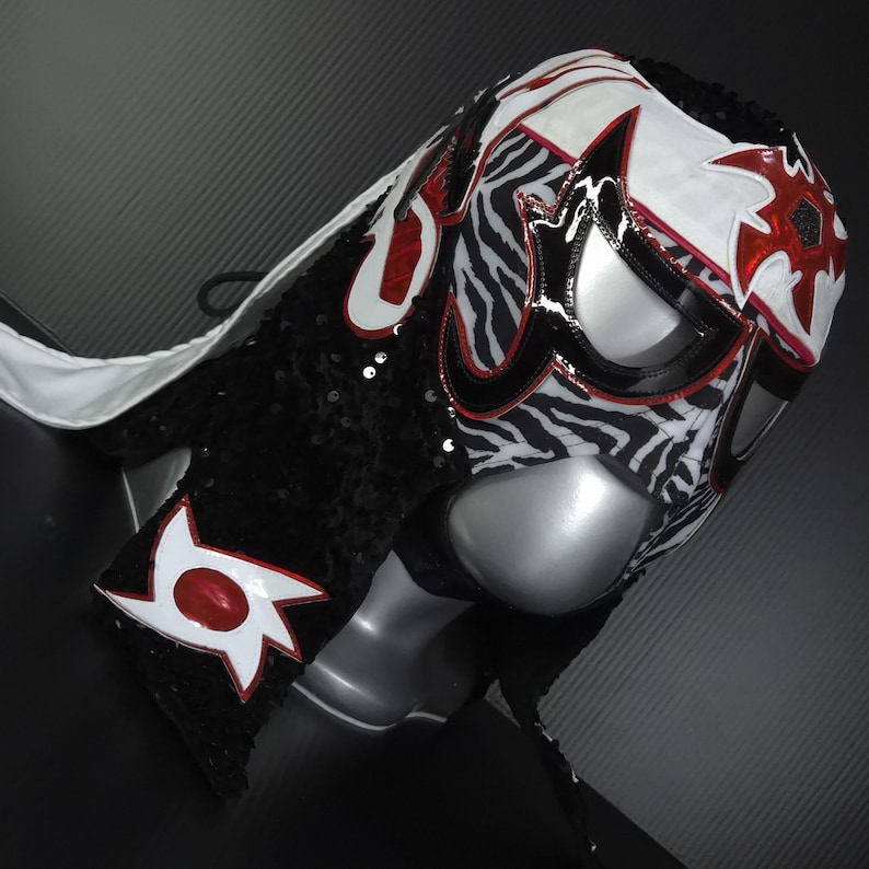 Puede incluir: Una m&aacute;scara de lucha libre con un dise&ntilde;o llamativo. La m&aacute;scara presenta un estampado de cebra en blanco y negro, acentuado con detalles rojos y blancos. Est&aacute; adornada con lentejuelas negras y una capa blanca.