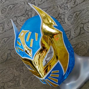 Op de afbeelding: Een blauw en gouden worstelmasker met een wit ontwerp. Het masker heeft een puntige bovenkant en een zilveren gaasooggebied.