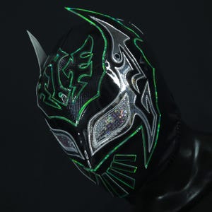 Op de afbeelding: Een zwart worstelmasker met groene en zilveren accenten. Het masker heeft een gestileerd ontwerp met een puntige bovenkant en een grote oogopening.
