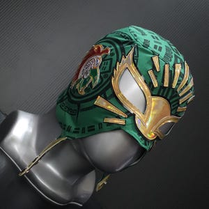 May include: A green and gold lucha libre wrestling mask. The mask features a gold and silver eye design, gold accents, and a circular emblem with a red and gold design. The mask is displayed on a mannequin head.