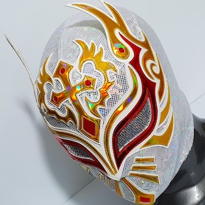 Op de afbeelding: Wit worstelmasker met gouden en rode ontwerpen. Het masker heeft een witte mesh-achtergrond met een gouden en rood tribale patroon. Het masker heeft een puntige bovenkant en een lange, vloeiende staart.