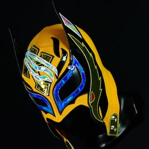 Peut inclure: Un masque de lutte jaune et noir avec des accents bleus et verts. Le masque a un motif de griffe sur le devant et un motif d'aile sur le côté.