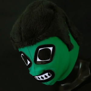 May include: Green wrestling mask with black and white details, featuring a large, stylized eye design and a toothy grin. The mask has a black fur-like hairpiece.