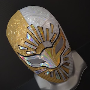 Op de afbeelding: Een goud en wit Lucha Libre worstelmasker. Het masker heeft een sunburst-ontwerp met gouden en zilveren accenten. De oog- en mondopeningen zijn zilverkleurig omlijnd. Het masker wordt gepresenteerd op een witte mannequinhoofd.