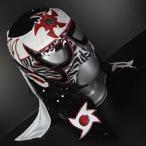 Puede incluir: Una m&aacute;scara de lucha libre con un dise&ntilde;o llamativo. La m&aacute;scara presenta un patr&oacute;n blanco y de cebra, acentuado con detalles rojos y negros. La m&aacute;scara est&aacute; adornada con lentejuelas y un dise&ntilde;o de estrella.