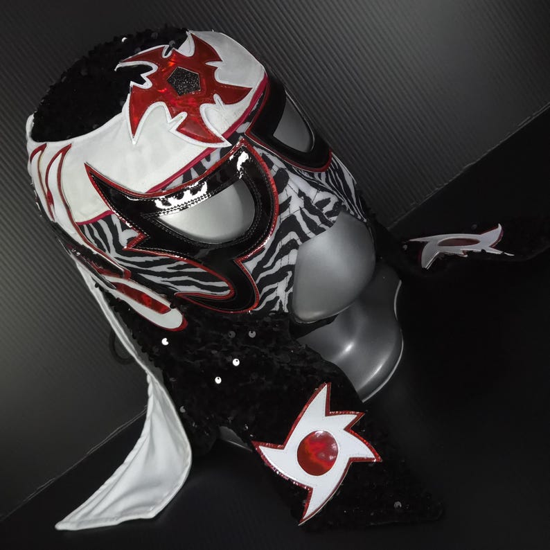 Puede incluir: Una m&aacute;scara de lucha libre con un dise&ntilde;o llamativo. La m&aacute;scara presenta un patr&oacute;n blanco y de cebra, acentuado con detalles rojos y negros. La m&aacute;scara est&aacute; adornada con lentejuelas y un dise&ntilde;o de estrella.