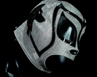 May include: A silver and black wrestling mask with a black design on the front. The mask has a black eye opening and a black mouth opening.