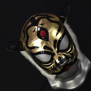 Puede incluir: Una máscara de lucha libre dorada y negra con diseño de tigre. La máscara presenta detalles negros, aberturas blancas para ojos y boca, y un detalle de ojo rojo. La máscara tiene orejas negras y está ribeteada con piel blanca.