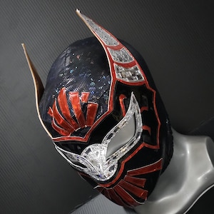 Puede incluir: Una máscara de lucha libre negra y roja con detalles plateados. La máscara presenta un diseño detallado con patrones rojos y plateados, incluyendo formas de alas y una tela negra texturizada. La máscara se muestra en una cabeza de maniquí.
