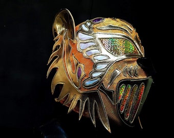 May include: A gold wrestling mask with a tiger design. The mask has a white and purple trim and iridescent sequins. The mask has a large, pointed ear on the left side.