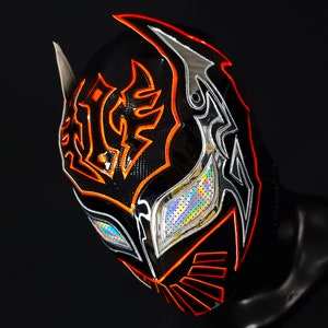 Op de afbeelding: Een zwart worstelmasker met oranje en zilveren accenten. Het masker heeft een gestileerd ontwerp met een grote, centrale oogopening en een kleinere, driehoekige opening op het voorhoofd.