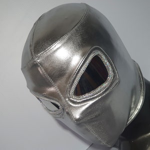Puede incluir: Una máscara de lucha libre plateada brillante con aberturas para los ojos adornadas con purpurina iridiscente. La máscara está hecha de un material liso y reflectante y está diseñada para cubrir toda la cara. Se muestra sobre una cabeza de maniquí negra.