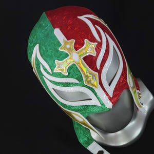 May include: A wrestling mask in red, white, and green. It has a gold cross design and white detailing around the eye and mouth. The mask is made from a shiny, textured fabric.