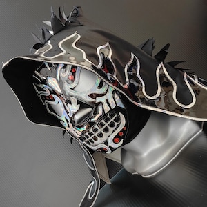 May include: A black and silver lucha libre mask with a hood. The mask has a skull design with red details and a silver flame pattern around the hood. The hood has black spikes.