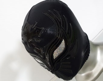 May include: Black wrestling mask with a detailed design featuring a cross and a stylised flame pattern. The mask has a shiny finish and rhinestones around the eye holes.