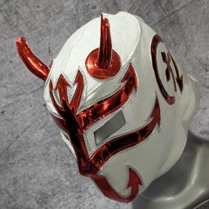 Puede incluir: Máscara de lucha libre blanca y roja con un diseño diabólico. La máscara presenta cuernos, flechas y detalles rojos, con un contorno plateado. Un emblema circular con una "X" está en el lateral. La máscara se muestra en una cabeza de maniquí.