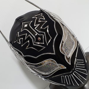 Op de afbeelding: Een zwart en zilveren worstelmasker met een geometrisch ontwerp. Het masker heeft een puntige bovenkant en een zilveren rand rond de ogen en de mond.