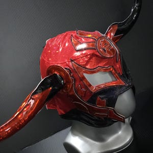 Peut inclure: Un masque de lutte rouge et noir avec une finition texturée et brillante. Le masque présente de grandes cornes incurvées et des détails complexes autour des ouvertures des yeux et de la bouche. Le design comprend un emblème circulaire sur le front.