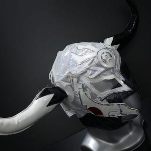 Peut inclure: Un masque de lutte argenté et noir avec une surface texturée et scintillante. Le masque présente un design cornu avec des cornes blanches et noires et un accent rouge. Le masque est présenté sur une tête de mannequin.
