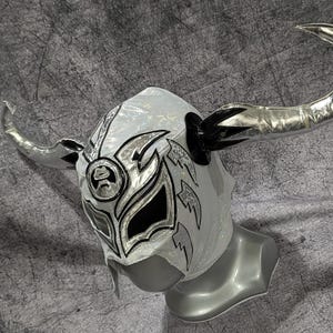 Peut inclure: Un masque de lutte argenté et noir doté de grandes cornes métalliques argentées incurvées sur les côtés. Le masque présente une base en tissu maillé texturé avec des motifs complexes en forme de flammes noires et argentées autour des yeux.