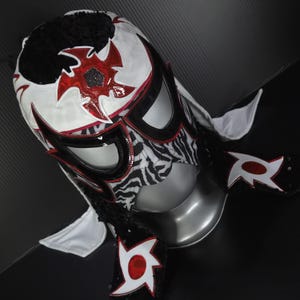 Puede incluir: Una m&aacute;scara de lucha libre profesional con una combinaci&oacute;n de colores blanco, negro y rojo. La m&aacute;scara presenta un patr&oacute;n de cebra en la cara, con detalles en rojo y un dise&ntilde;o de estrella. La parte superior de la m&aacute;scara es negra con una textura esponjosa.
