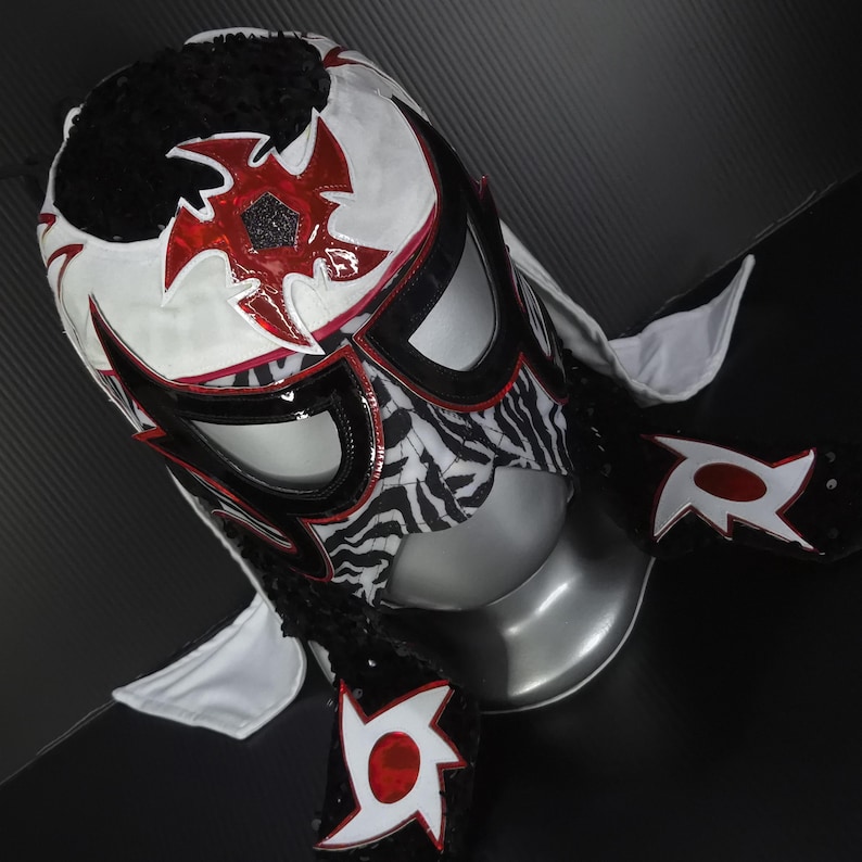 Puede incluir: Una m&aacute;scara de lucha libre profesional con una combinaci&oacute;n de colores blanco, negro y rojo. La m&aacute;scara presenta un patr&oacute;n de cebra en la cara, con detalles en rojo y un dise&ntilde;o de estrella. La parte superior de la m&aacute;scara es negra con una textura esponjosa.