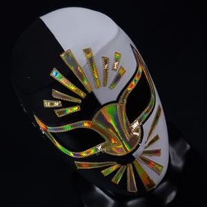 Op de afbeelding: Een zwart-wit worstelmasker met gouden en iriserende accenten. Het masker heeft een gestileerd ontwerp met een zonnestralenpatroon en een grote oogopening.
