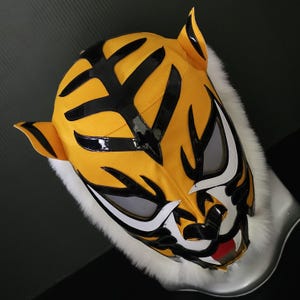 Może przedstawiać: Żółto-czarna maska tygrysa z białym futrzanym wykończeniem. Maska ma czarne paski, białe akcenty wokół oczu i ust oraz czerwony język. Maska ma przypominać twarz tygrysa.