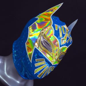 Op de afbeelding: Een blauw lucha libre masker met iriserende accenten en witte rand. Het masker heeft een gedetailleerd ontwerp met gouden en zilveren versieringen en twee witte hoorns. Het masker is gemaakt van een glanzend, getextureerd materiaal.