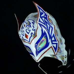 May include: A white lucha libre mask with blue and red accents. The mask features a glossy finish with intricate designs, including geometric shapes and stylized patterns. The mask has two pointed ear-like extensions and a chin strap.