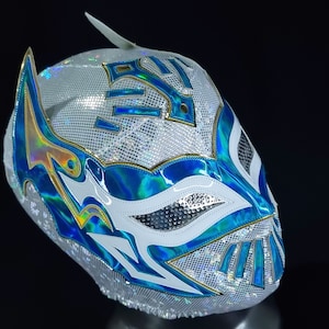 May include: A white wrestling mask with blue and gold accents. The mask has a geometric design and a pointed top.