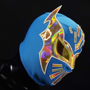 Op de afbeelding: Een levendig blauw worstelmasker met gouden en iriserende accenten. Het masker heeft een gestileerd ontwerp met scherpe hoeken en decoratieve elementen. De gouden rand omlijnt de oog- en mondopeningen, wat een opvallend contrast creëert.