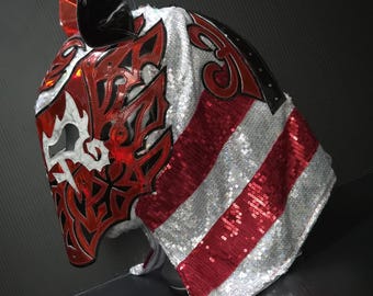 May include: A professional wrestling mask with a red and black design. The mask features a stylized wolf or dog face with intricate cutouts and a sequined silver and red striped cape. The mask has a glossy black horn.