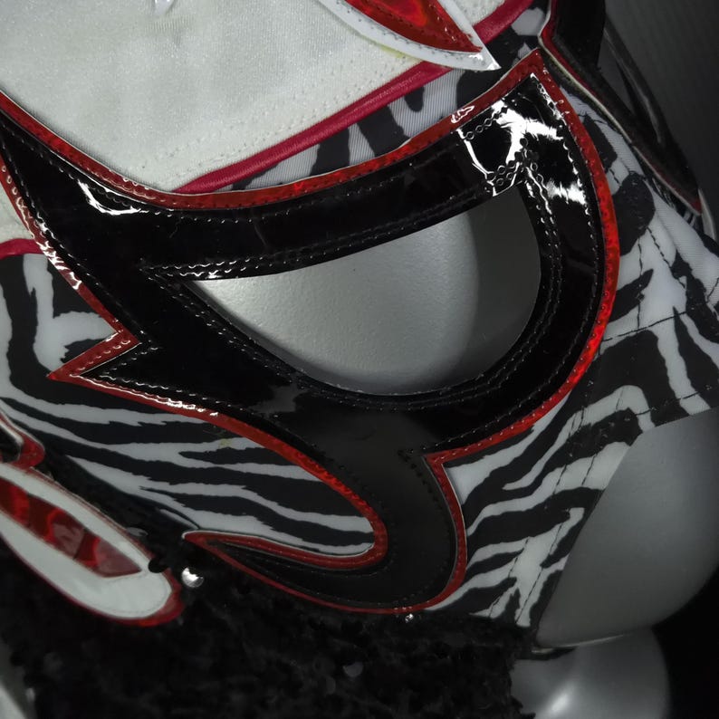 Puede incluir: Primer plano de una m&aacute;scara de lucha libre con dise&ntilde;o de estampado de cebra. La m&aacute;scara presenta detalles en negro y rojo, con un acabado brillante. Las aberturas de los ojos est&aacute;n delineadas en negro y la parte superior de la m&aacute;scara es blanca.
