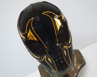 May include: A black wrestling mask with gold accents and a mesh design. The mask has a pointed top and a wide, open mouth.