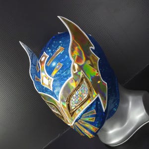 Op de afbeelding: Een blauw en goud lucha libre masker met een glinsterend, iriserend ontwerp. Het masker heeft een gedetailleerd patroon met gouden accenten en witte randen. Het masker wordt getoond op een mannequinhoofd.