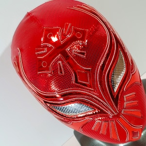 Op de afbeelding: Een rood worstelmasker met een wit ontwerp. Het masker heeft een metallic glans en een gedetailleerd patroon.