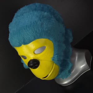May include: A blue and yellow monkey mask with a black nose. The mask has a yellow face with eye holes and a black nose. The top of the mask is covered in blue fur. The mask is displayed on a gray mannequin head.