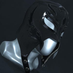 Op de afbeelding: Een zwart worstelmasker met glanzende zwarte accenten en een vlamachtig ontwerp. Het masker heeft oog- en mondopeningen en wordt vastgezet met drukknopen. Het masker wordt getoond op een mannequinhoofd.