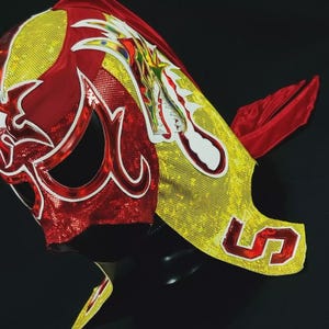 Op de afbeelding: Een rood en geel worstelmasker met een drakenontwerp en het nummer "5" aan de zijkant. Het masker heeft een witte rand en een rode cape.