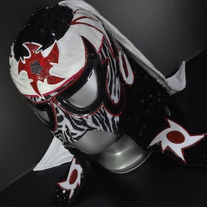 Puede incluir: Una m&aacute;scara de lucha libre con un dise&ntilde;o llamativo. La m&aacute;scara presenta una base blanca con detalles rojos y negros, incluido un estampado de cebra en la cara. Est&aacute; adornada con lentejuelas y una capucha blanca.